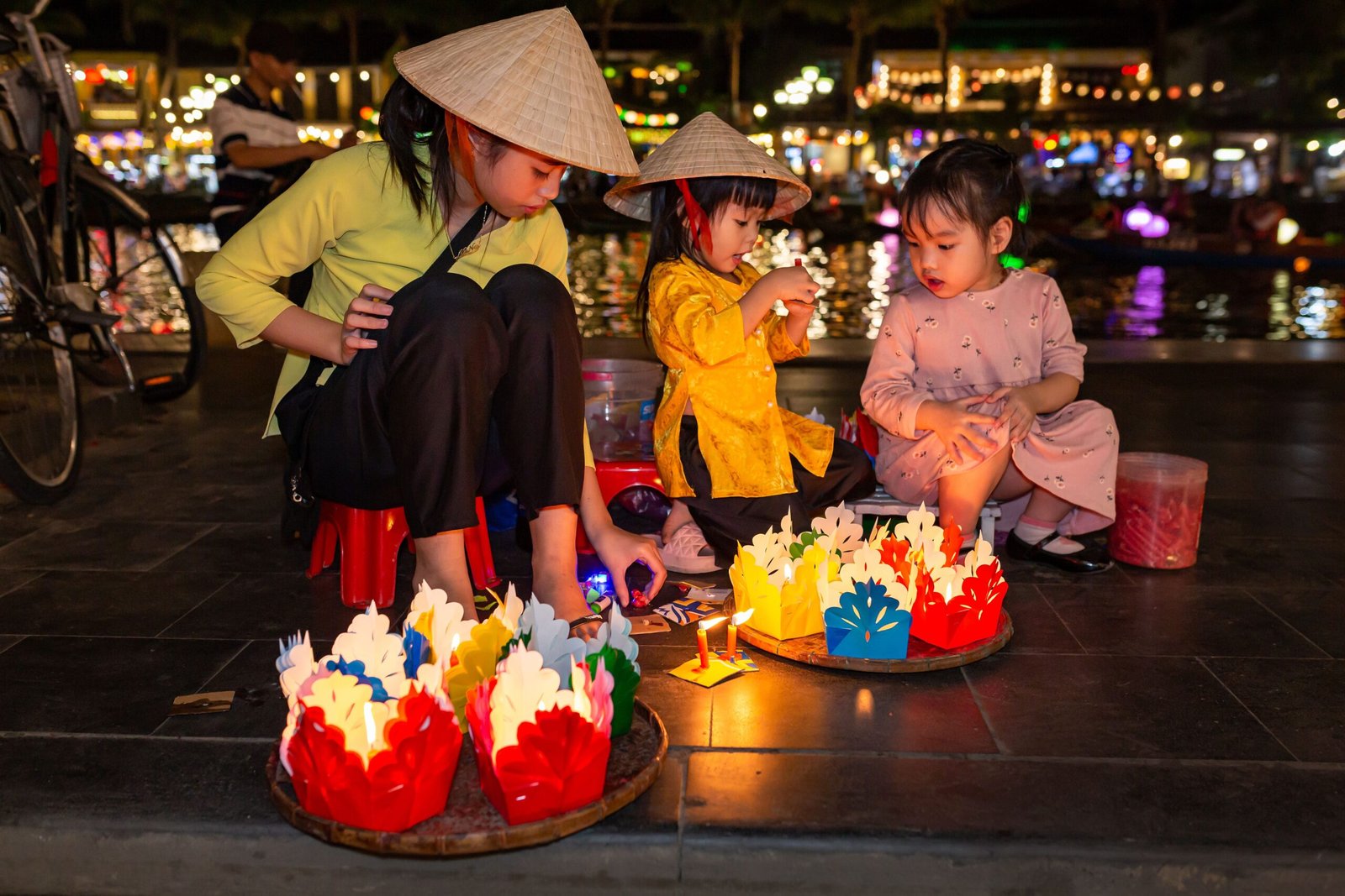 Two young children in conical hats making paper lanterns by the river in Hoi An at night